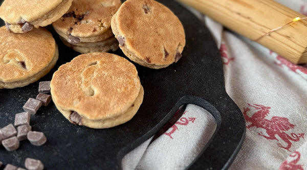 Chocolate Chip Welsh Cakes on a bakestone griddle with a rolling pin, chocolate chunks and a welsh dragon table cloth