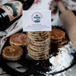 Load image into Gallery viewer, Packaged Barti Spiced Rum and Raisin Welsh Cakes on a bakestone surrounded by welsh cakes, ingredients, rolling pin and barti spiced rum bottle
