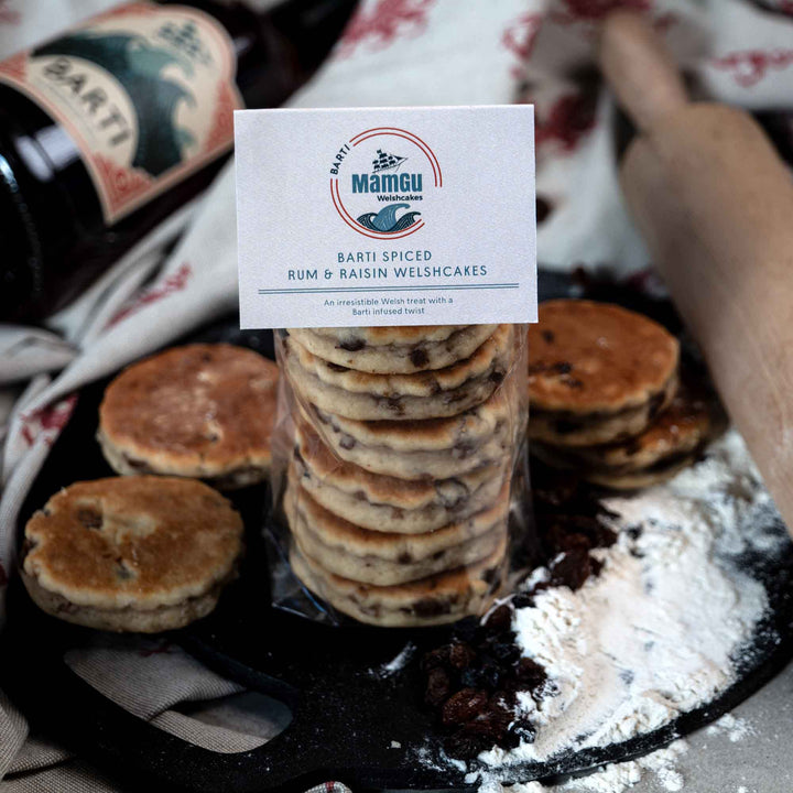 Packaged Barti Spiced Rum and Raisin Welsh Cakes on a bakestone surrounded by welsh cakes, ingredients, rolling pin and barti spiced rum bottle