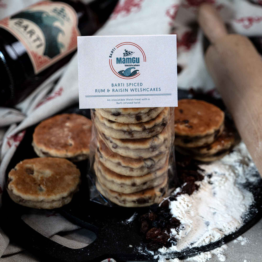 Packaged Barti Spiced Rum and Raisin Welsh Cakes on a bakestone surrounded by welsh cakes, ingredients, rolling pin and barti spiced rum bottle