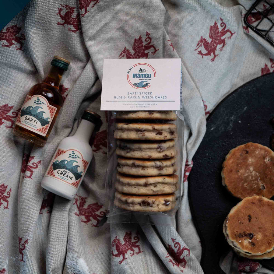 Packaged Barti Spiced Rum and Raisin Welsh Cakes on a welsh dragon table cloth next to welsh cakes and miniature bottles of barti rum and barti cream. 