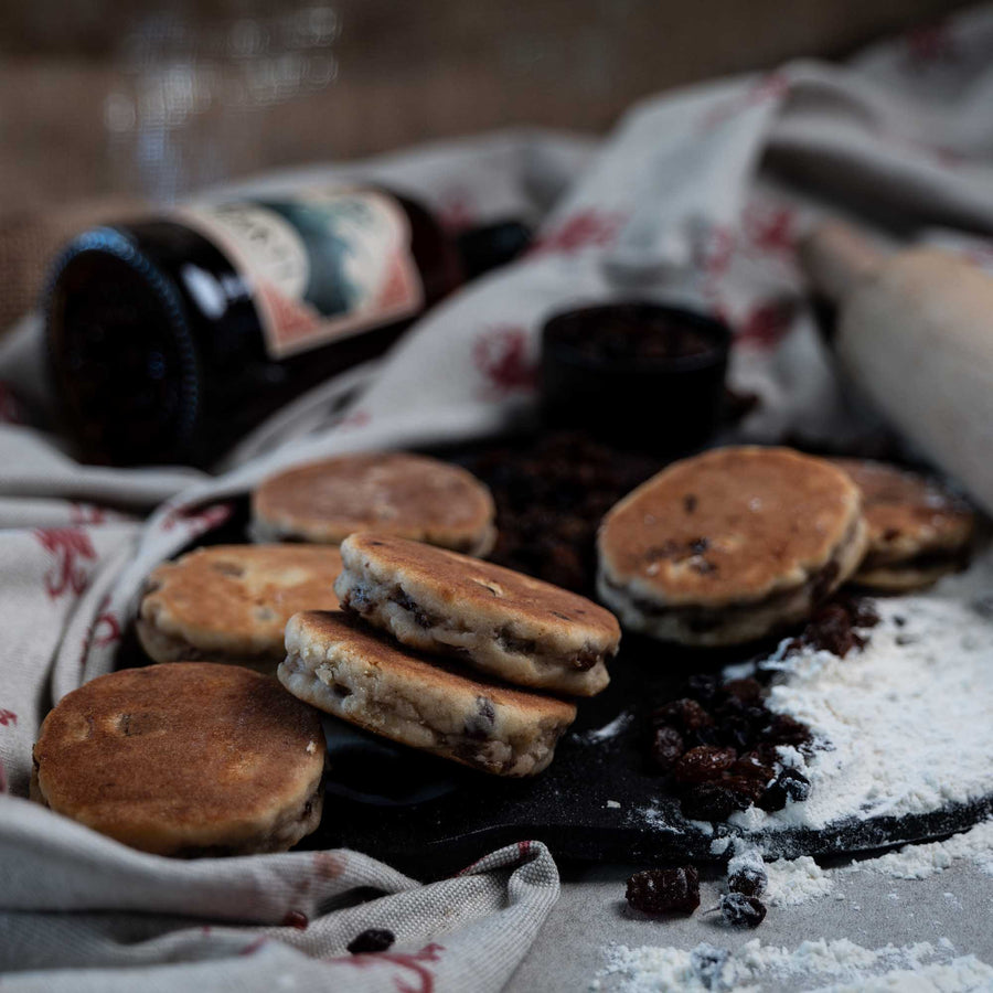 Barti Spiced Rum and Raisin Welsh Cakes on a bakestone surrounded by ingredients, rolling pin and barti spiced rum bottle