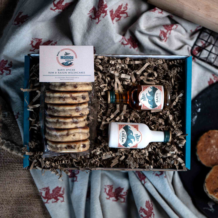 Packaged Barti Spiced Rum & Raisin Welsh Cakes with a miniature Barti Spiced Rum bottle and a miniature Barti Cream bottle in a gift box on a table cloth with welsh dragons. 