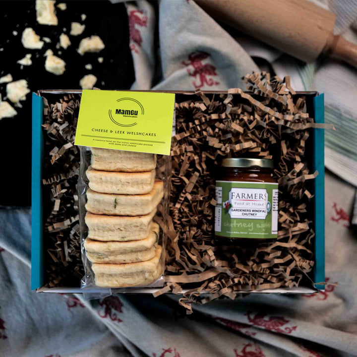 Packaged Cheese & Leek Welsh Cakes with a jar of chutney in a gift box displayed on a messy kitchen work top with rolling pin, griddle and welsh dragon table cloth.