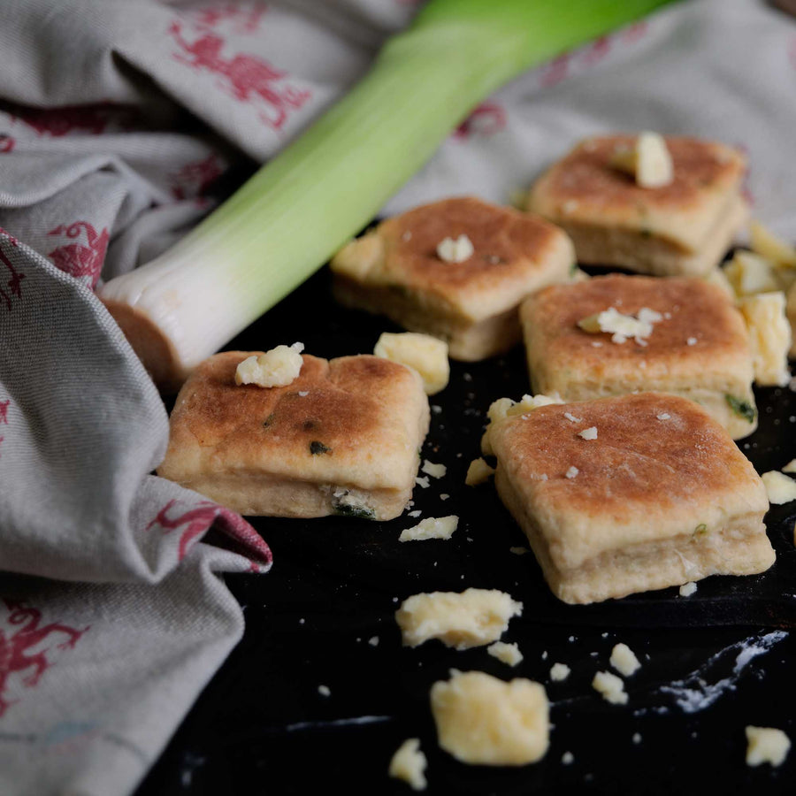 Square Cheese and Leek Welsh Cakes on a griddle with broken cheese and a large leek.