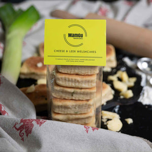 Packaged Cheese and Leek Welsh Cakes with a green MamGu Welshcakes label on a busy background with leeks, cheese and kitchen utensils. 