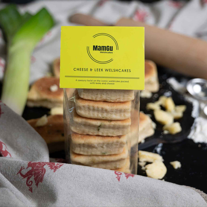 Packaged Cheese and Leek Welsh Cakes with a green MamGu Welshcakes label on a busy background with leeks, cheese and kitchen utensils. 