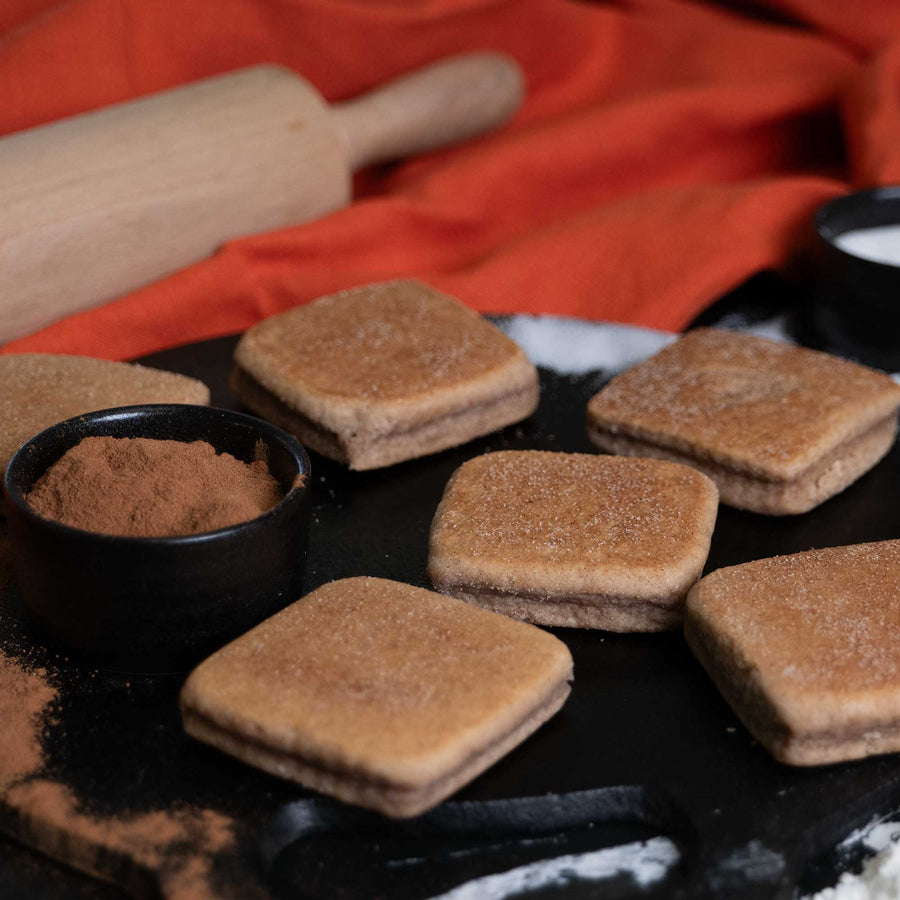 Square Cinnamon Welsh Cakes on a black griddle/bakestone next to a ramekin of cinnamon and a rolling pin.