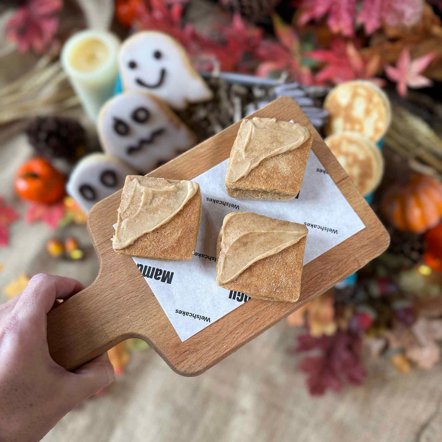 Three Chai flavoured Welsh Cakes with triangle icing on a wooden board with Halloween-themed decorations in the background.