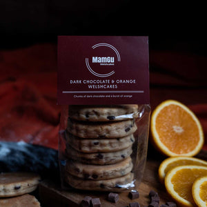 Packaged dark chocolate orange Welshcakes with oranges and chocolate pieces on a wooden board.