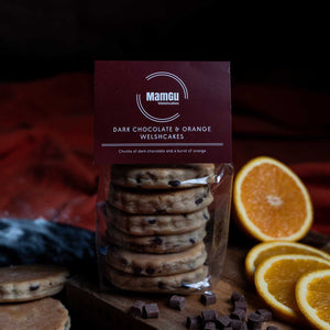 Packaged dark chocolate orange Welshcakes with oranges and chocolate pieces on a wooden board.