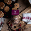 Load image into Gallery viewer, Dark Chocolate &amp; Raspberry Welshcakes in a pack with a purple label on a messy rustic worktop with loose raspberry and chocolate chips
