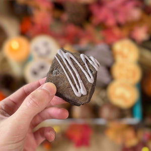 Hand holding a Black Velvet Chocolate Welsh Cake with Purple icing against a blurred background of Autumn decorations.