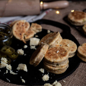 stack of La Mexicana Welsh Cakes on a black griddle backstone with cheese and jalapeños scattered around.