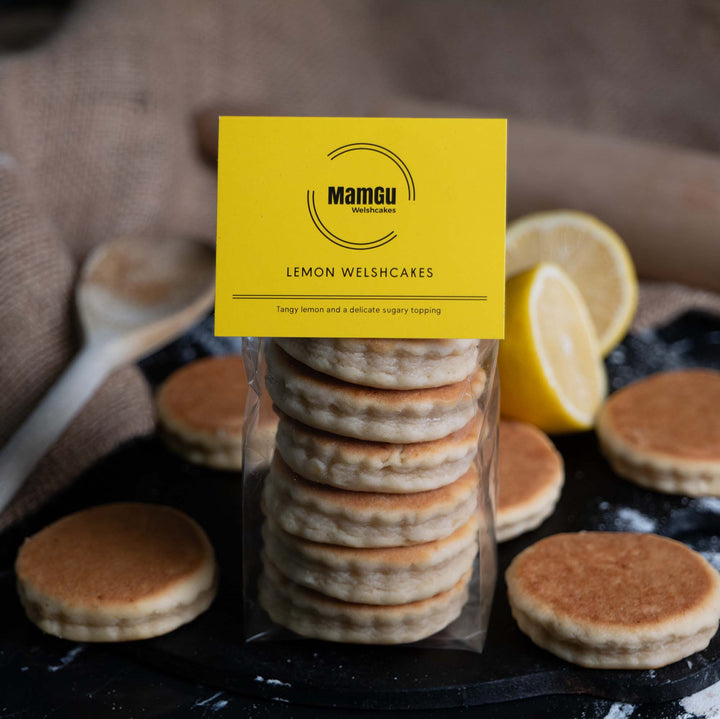 Packaged lemon Welshcakes with a yellow MamGu Welshcakes label with a rustic background of welsh cakes, lemons, griddle bakestone and wooden spoon.