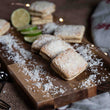 Load image into Gallery viewer, Lime and Coconut Welsh Cakes on a rustic kitchen work top on a wooden chopping board with loose desiccated coconut and sliced lime.
