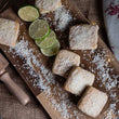 Load image into Gallery viewer, Lime and Coconut Welsh Cakes on a rustic kitchen work top on a wooden chopping board with loose desiccated coconut and sliced lime.
