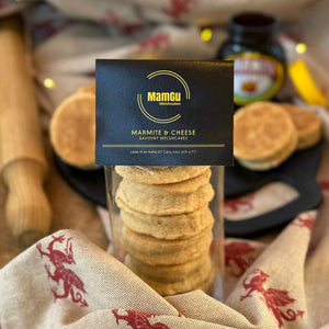 6 marmite welsh cakes in clear packaging with a black mamgu welshcakes label on top. it is on top of a messy kitchen work top with a rolling pin to the left, a black bakestone in the background, loose welsh cakes and an open jar of marmite.