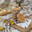 Load image into Gallery viewer, Mini Egg Welsh Cakes stacked on top of each other on a wooden chopping board. The table has a table cloth with welsh dragons on it and in the background is loose mini eggs and a wooden rolling pin.
