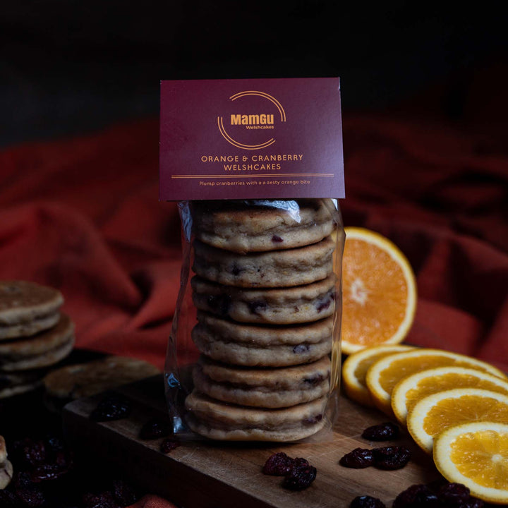 Packaged Orange and Cranberry Welsh Cakes on a wooden chopping board with sliced orange and loose cranberries