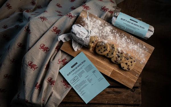 A styled flat-lay of a welsh cake dough gift set on a rustic wooden table. The image includes a wooden board with welsh cake dough cut up, dusting of flour, a rolled packaged dough, and a printed recipe card, all arranged next to a welsh dragon patterned beige and red table cloth.