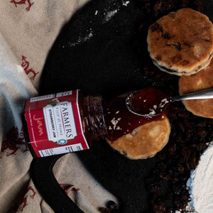 Strawberry jam being spread on traditional welsh cakes with a jar of jam, griddle and a ingredients visible.