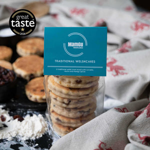 Packaged Traditional Welsh Cakes with a MamGu Welshcakes label on busy kitchen background with welsh dragon table cloth and welsh cakes on a griddle. Great Taste Award 1 gold star badge in the top left corner of the image. 