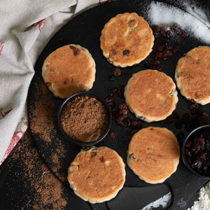 Welsh Cakes on a griddle with ingredients, spices, sugar, raisins on the side.