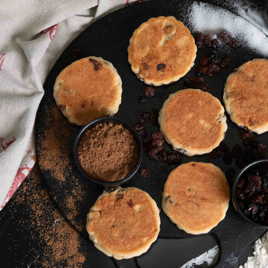 Welsh Cakes on a griddle with ingredients, spices, sugar, raisins on the side.