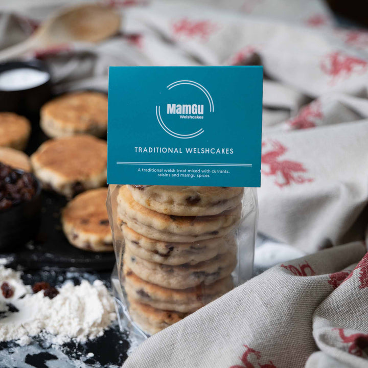 Packaged Traditional Welsh Cakes with a MamGu Welshcakes label on busy kitchen background with welsh dragon table cloth and welsh cakes on a griddle