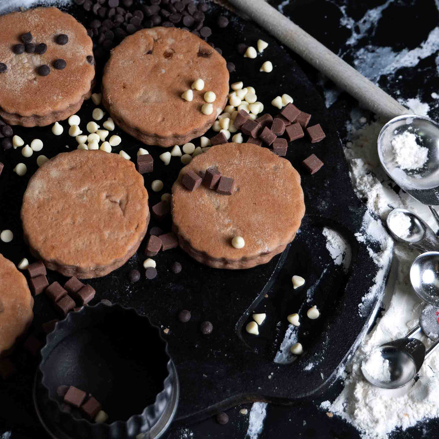 Triple Chocolate Welsh Cakes on a messy griddle with chocolate chips scattered all around, flour and a welsh cake cutter.