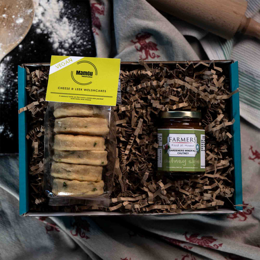 Packaged Vegan Cheese & Leek Welsh Cakes with a jar of chutney in a gift box displayed on a messy kitchen work top with rolling pin, griddle and welsh dragon table cloth.