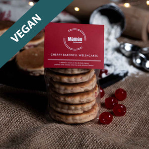 Packaged Cherry Bakewell Welsh Cakes with a MamGu Welshcakes Red label with loose welsh cakes, rolling pin, flour and fairy lights in the background.