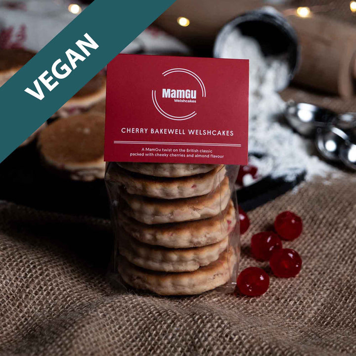 Packaged Cherry Bakewell Welsh Cakes with a MamGu Welshcakes Red label with loose welsh cakes, rolling pin, flour and fairy lights in the background.
