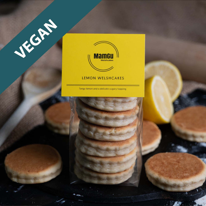 Packaged lemon Welshcakes with a yellow MamGu Welshcakes label with a rustic background of welsh cakes, lemons, griddle bakestone and wooden spoon.