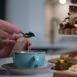 Person adding tea to a teabag next to a blue teacup with a blurred background of snacks.
