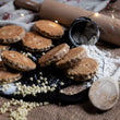 Load image into Gallery viewer, White chocolate chip welsh cakes Loose on a black bakestone, sitting on a messy rustic kitchen worktop surrounded by ingredients and wooden cutlery
