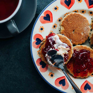 Plate with traditional welsh cakes topped with jam and clotted cream, next to a cup of tea on a dark surface.