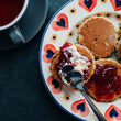 Load image into Gallery viewer, Plate with traditional welsh cakes topped with jam and clotted cream, next to a cup of tea on a dark surface.
