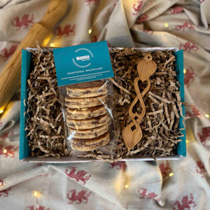 Welsh Love Spoon and Traditional Welsh Cakes in a gift box on top of a welsh dragon table cloth (beige with little red dragons) and in the blurred background is a wooden rolling pin and fair lights.