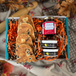 Load image into Gallery viewer, A gift box filled with orange shredded paper, containing three ghost shaped welsh cakes, a jar of Farmers' Food at Home Strawberry Jam, with two small Daffodil Foods Clotted Scream, photographed on a patterned fabric background with autumn leaves.
